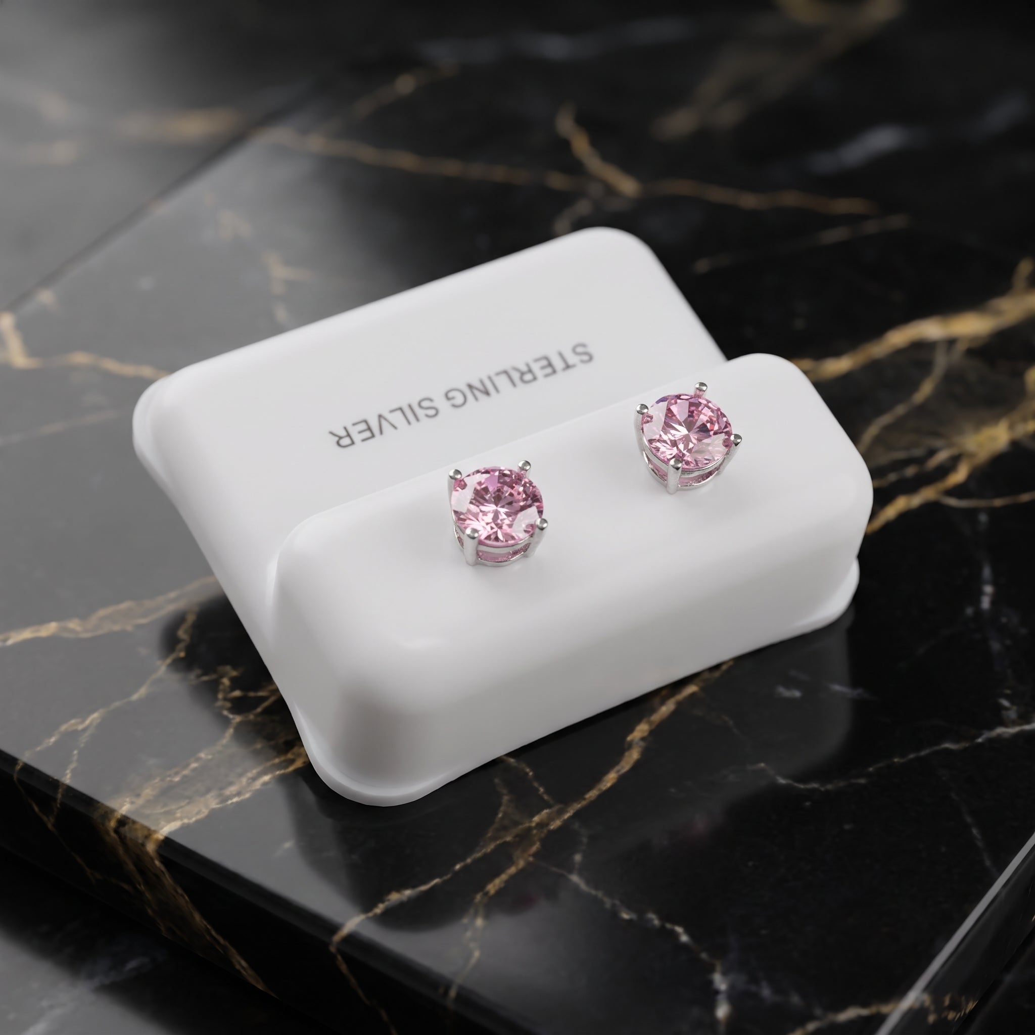 A pair of silver stud earrings with round pink gemstones, displayed on a white jewelry stand over a black marble surface with gold veining.