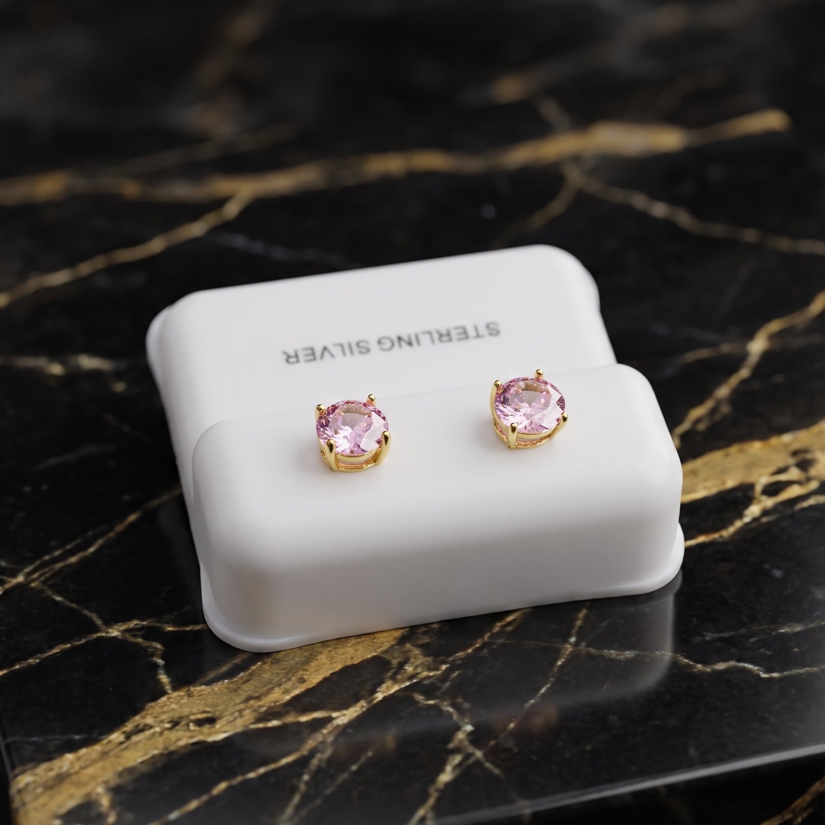 A pair of stud earrings with round pink gemstones in gold settings, displayed on a white jewelry stand labeled 'STERLING SILVER' on a black marble surface with gold veining.