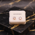 A pair of rose gold stud earrings with clear round-cut gemstones, presented on a cream-colored 'STERLING SILVER' display card, set against a black and gold marble background.