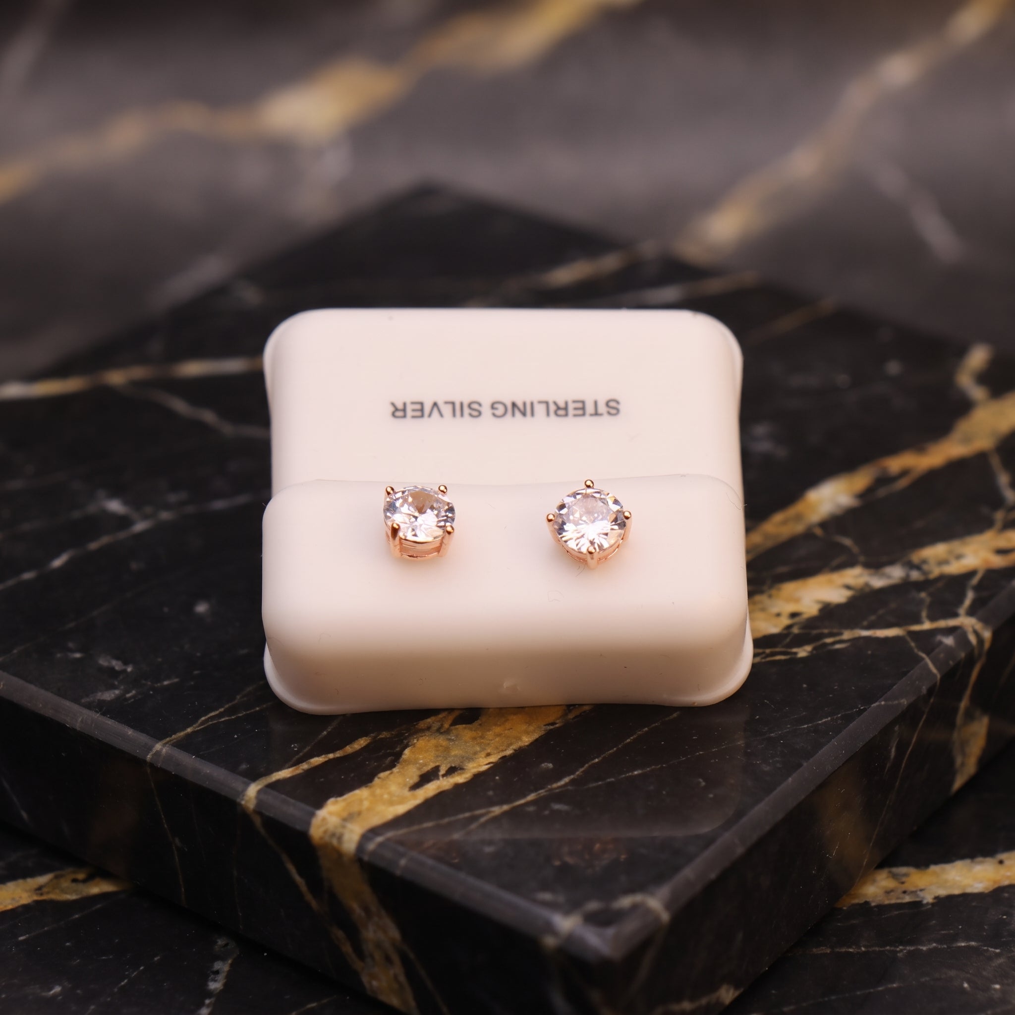 A pair of rose gold stud earrings with clear round-cut gemstones, presented on a cream-colored 'STERLING SILVER' display card, set against a black and gold marble background.