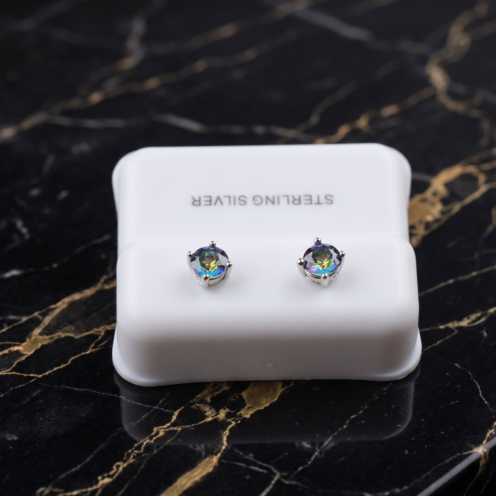 A pair of silver stud earrings with iridescent blue-green gemstones, displayed on a white 'STERLING SILVER' jewelry box set on a black marble surface with gold veining. mystic blue
