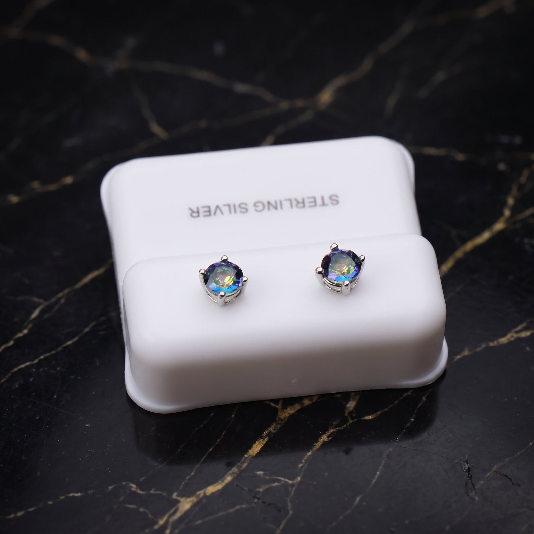 A pair of silver stud earrings with round iridescent blue-green stones, displayed on a white jewelry stand labeled 'STERLING SILVER' on a black marble surface.