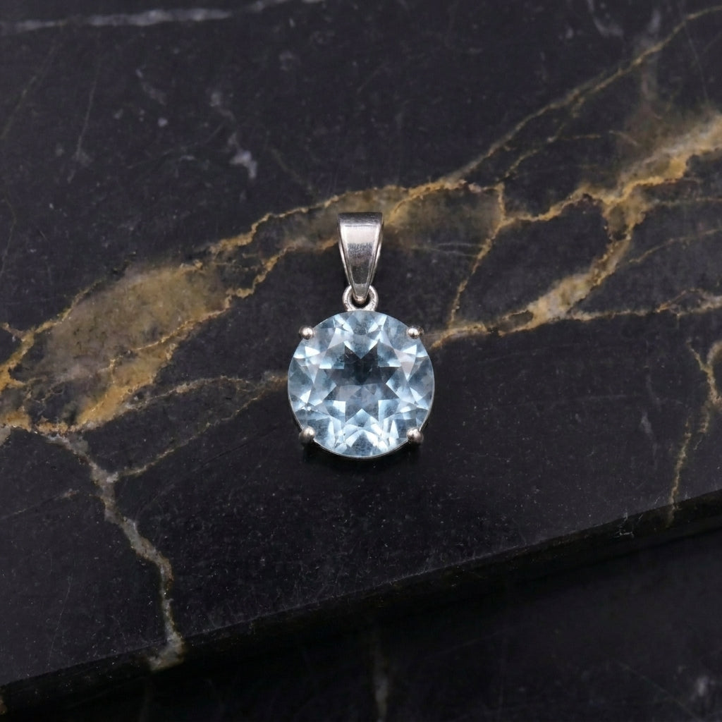 A round-cut light blue gemstone pendant in a silver-toned setting, displayed on a black and gold veined marble surface.