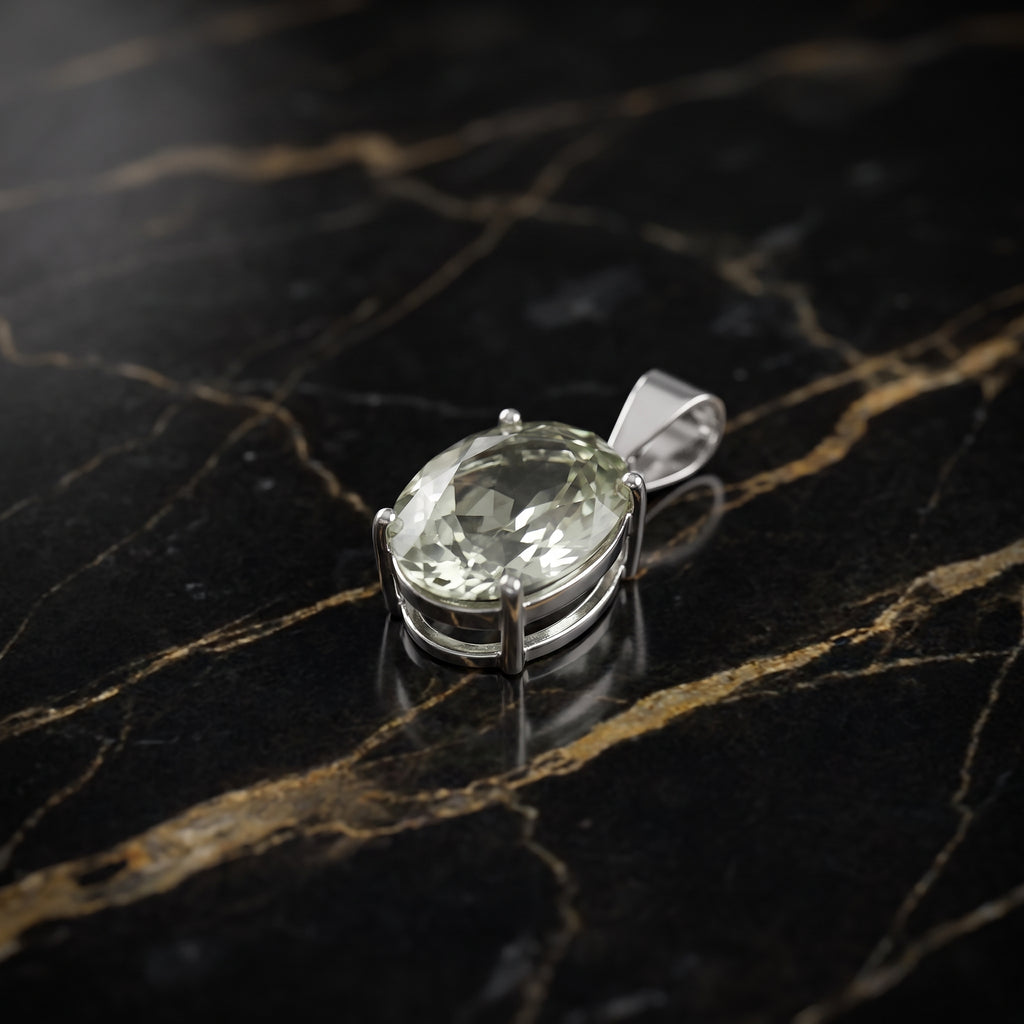 An oval-cut clear gemstone pendant in a silver-toned setting, photographed on a polished black marble surface with gold veining.