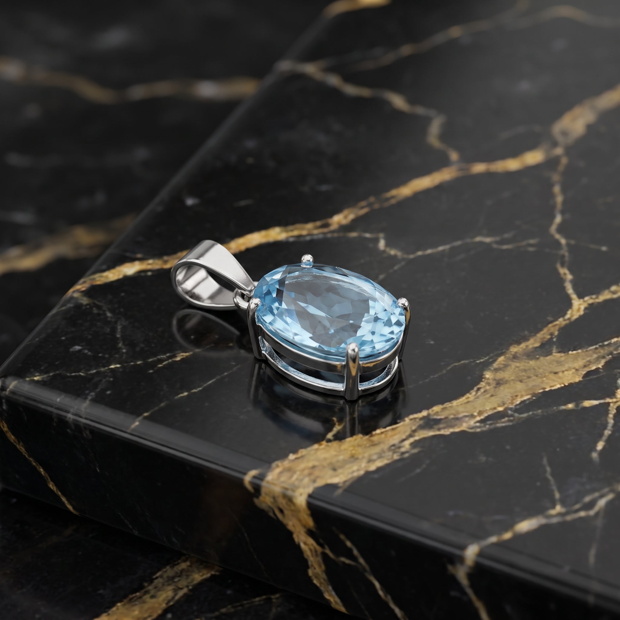 An oval-cut light blue gemstone pendant in a silver-toned setting, displayed on a black marble slab with gold veining.