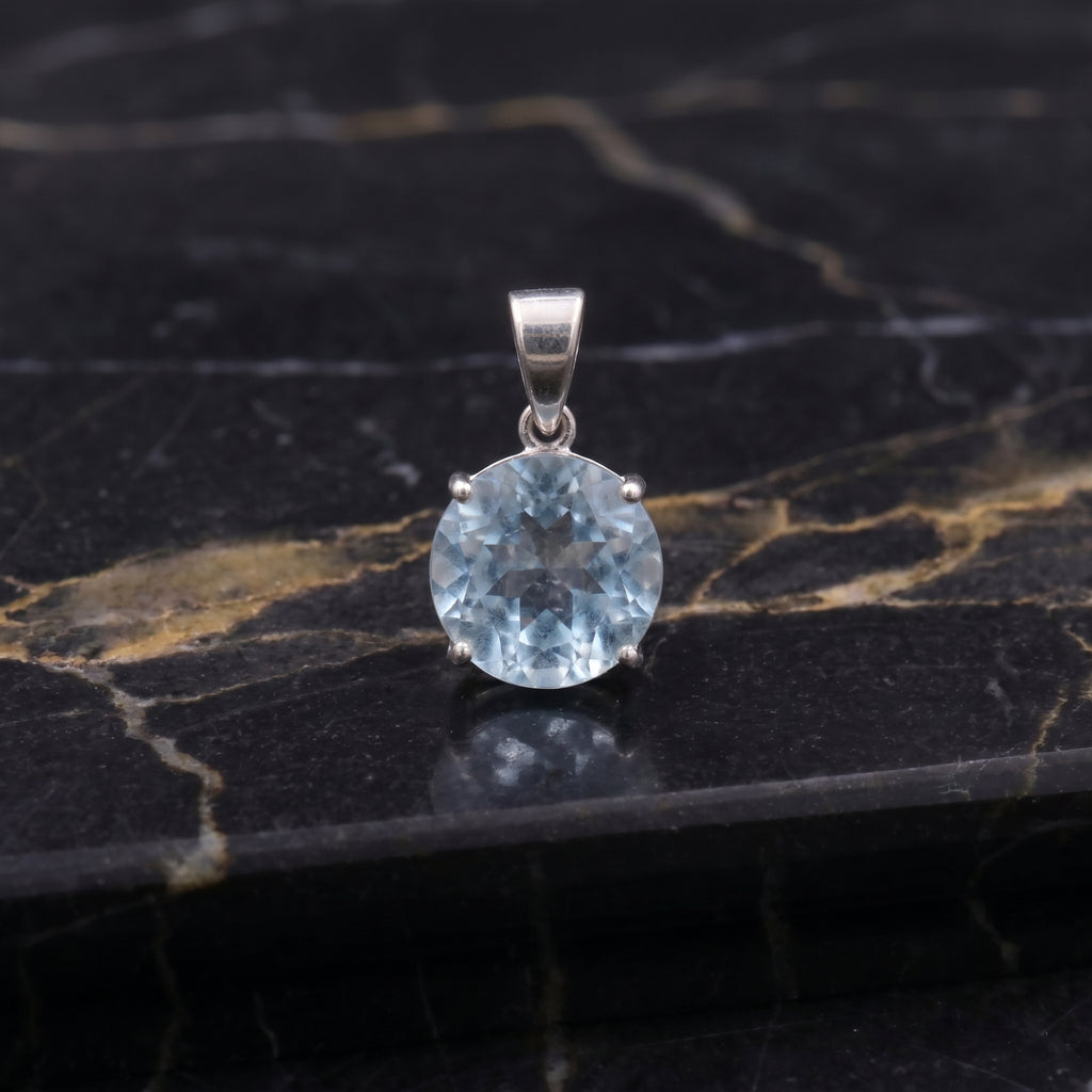 A round-cut light blue gemstone pendant in a silver-toned setting, displayed on a polished black marble surface with gold veining.