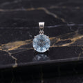 A round-cut light blue gemstone pendant in a silver-toned setting, displayed on a polished black marble surface with gold veining.