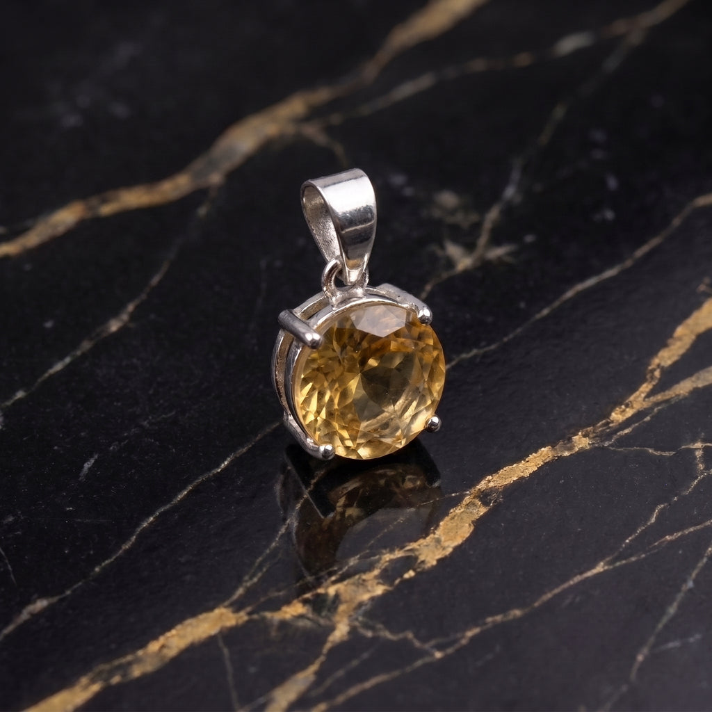 A round-cut yellow gemstone pendant in a silver-toned setting, displayed on a black marble surface with gold veining.