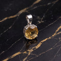 A round-cut yellow gemstone pendant in a silver-toned setting, displayed on a black marble surface with gold veining.