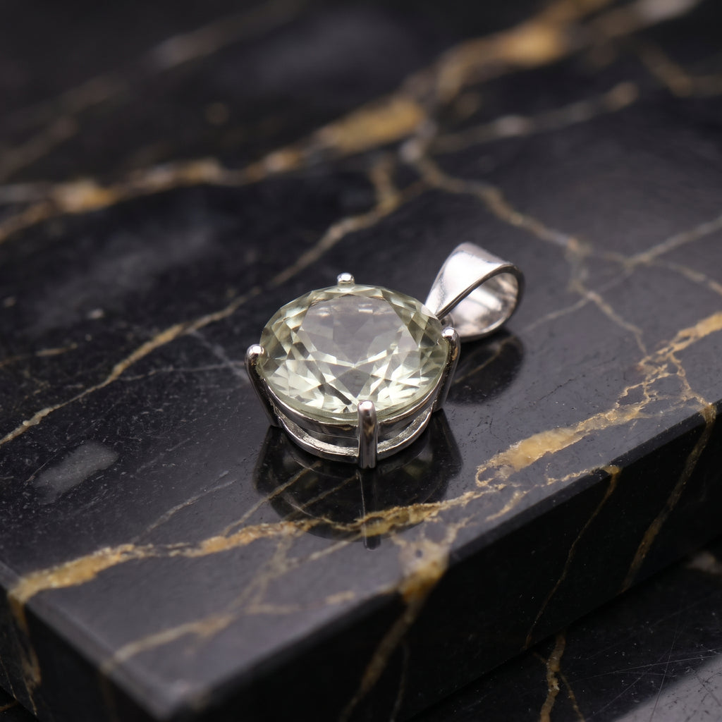 A silver pendant with a large, round, faceted pale green gemstone, resting on a polished black and gold veined marble slab.