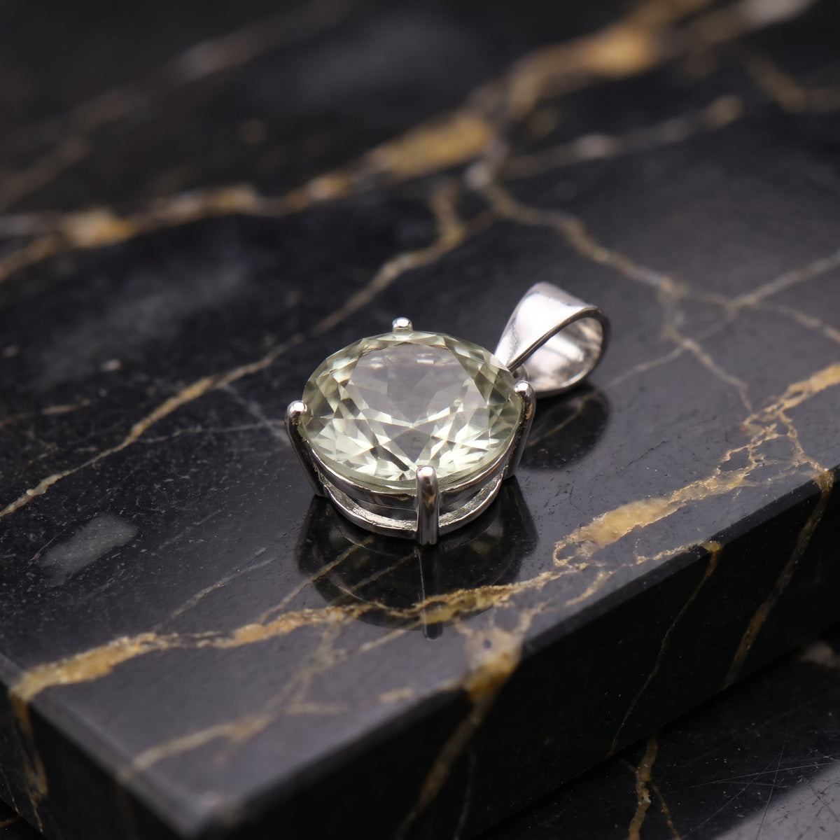 A silver pendant with a large, round, faceted pale green gemstone, resting on a polished black and gold veined marble slab.