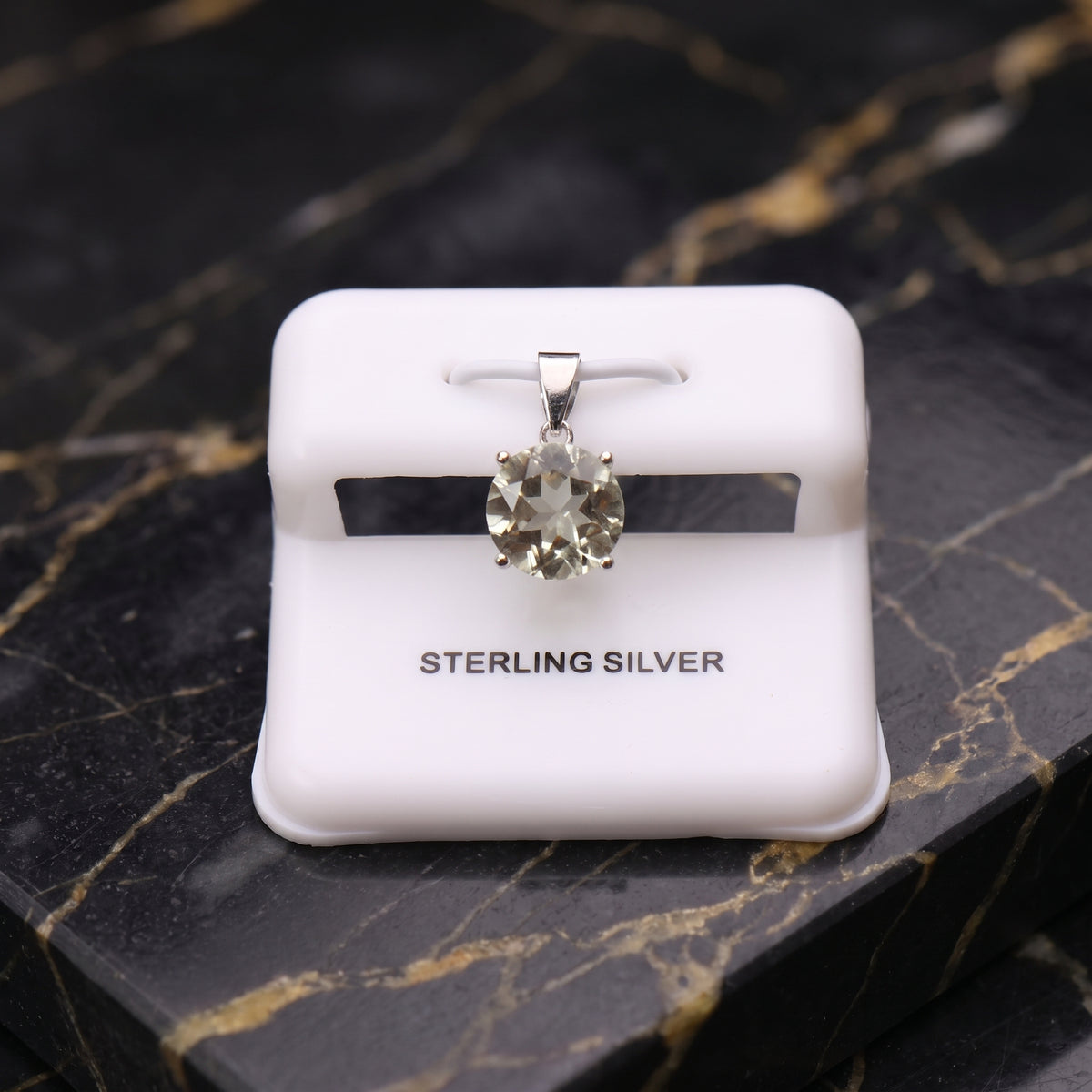 A sterling silver solitaire pendant with a clear oval-cut gemstone, displayed on a white jewelry stand labeled 'STERLING SILVER' against a black and gold veined marble background.
