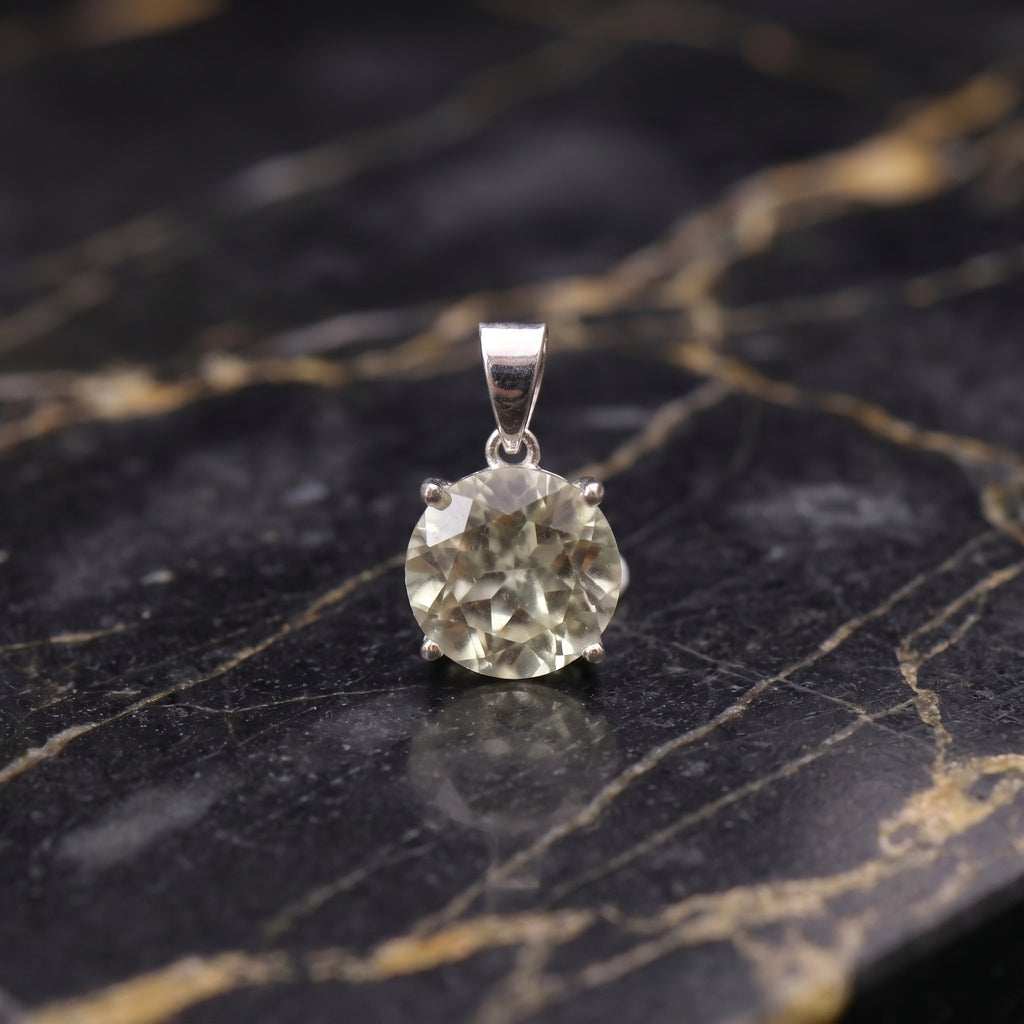 A round-cut pale yellow gemstone pendant set in a silver-toned metal prong, displayed on a black and gold veined marble surface.