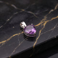 A heart-shaped purple gemstone pendant in a silver setting, displayed on a black marble surface with gold veining.