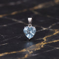 A heart-shaped light blue gemstone pendant in a silver setting, displayed on a black and gold veined marble surface.