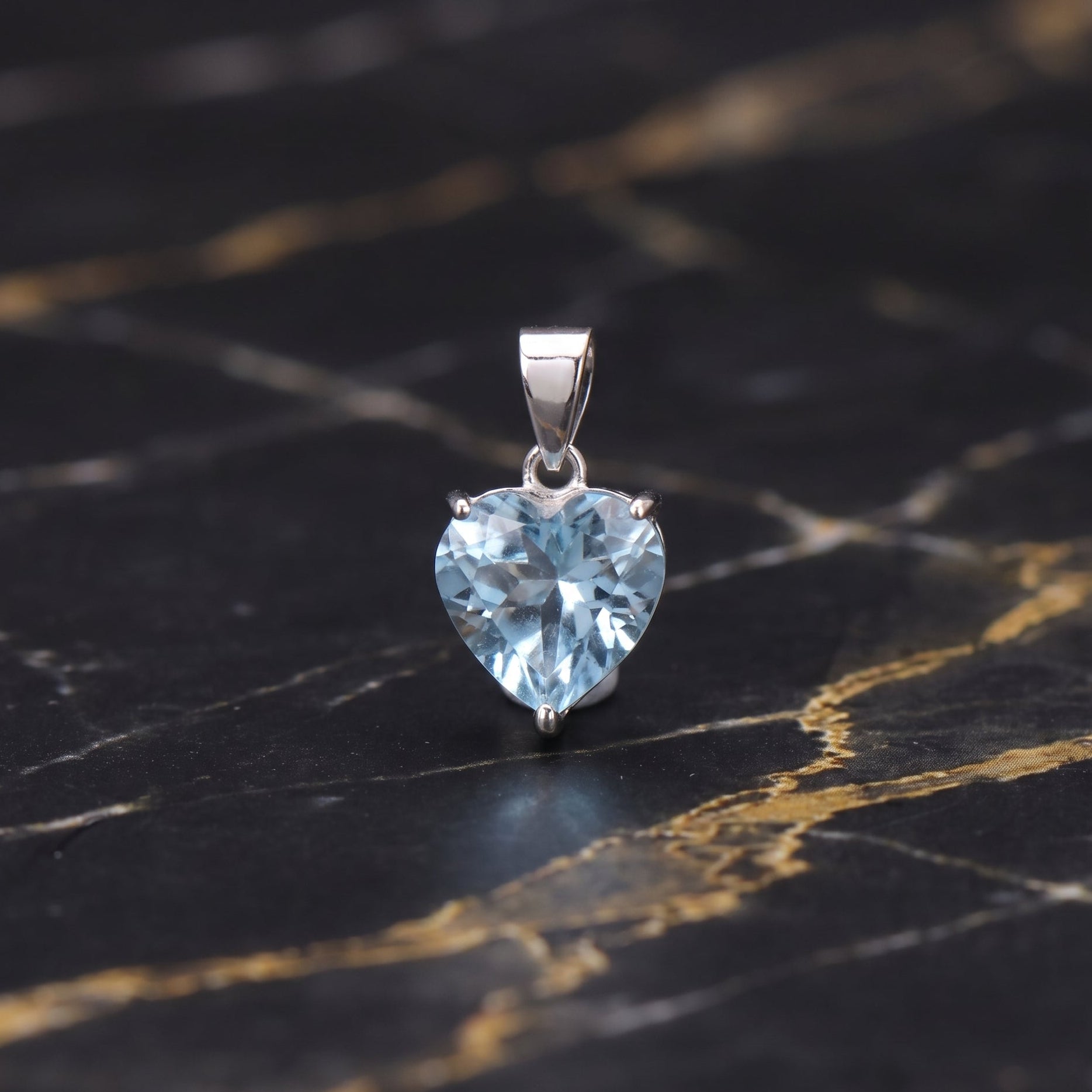 A heart-shaped light blue gemstone pendant in a silver setting, displayed on a black and gold veined marble surface.