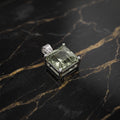 A square-cut green gemstone pendant in a silver-toned metal setting, displayed on a black marble surface with gold veining.