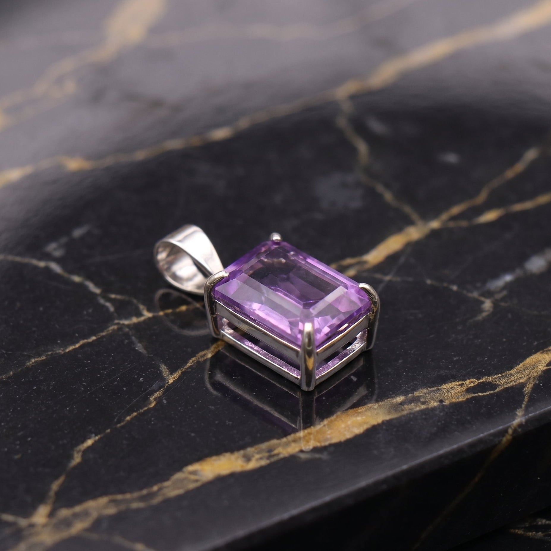 A rectangular purple gemstone pendant in a silver-toned metal setting, displayed on a black marble surface with gold veining.