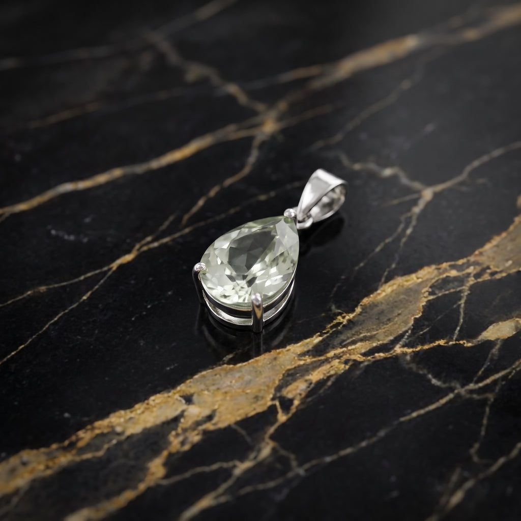 A teardrop-shaped clear gemstone pendant in a silver-colored metal setting, displayed on a black marble surface with gold veining.