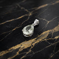 A teardrop-shaped clear gemstone pendant in a silver-colored metal setting, displayed on a black marble surface with gold veining.