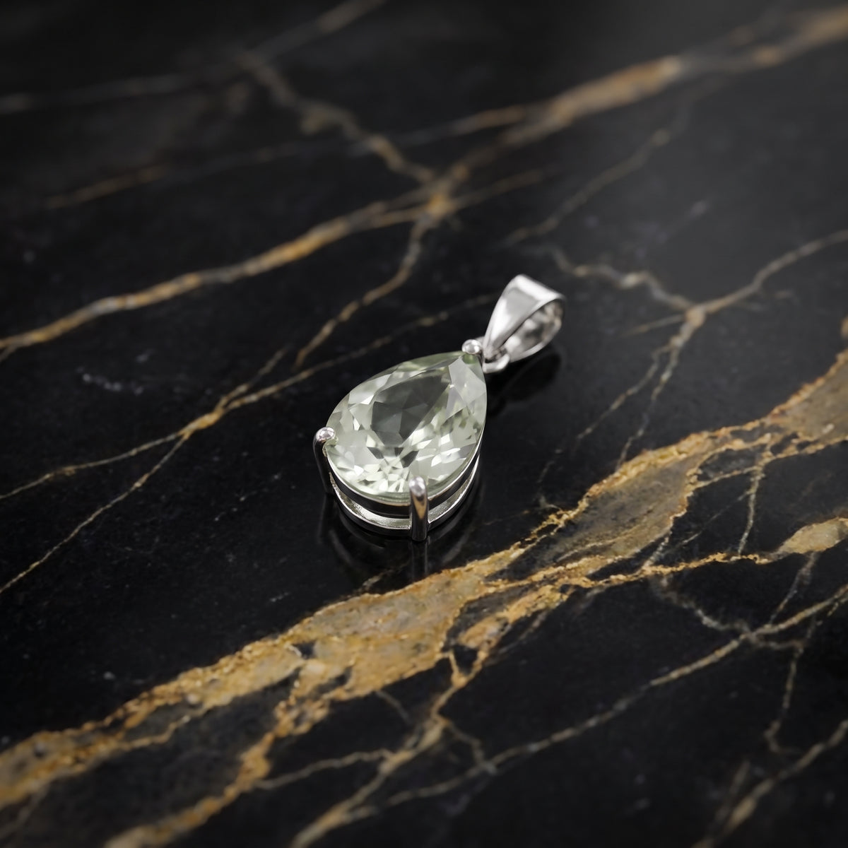 A teardrop-shaped clear gemstone pendant in a silver-colored metal setting, displayed on a black marble surface with gold veining.