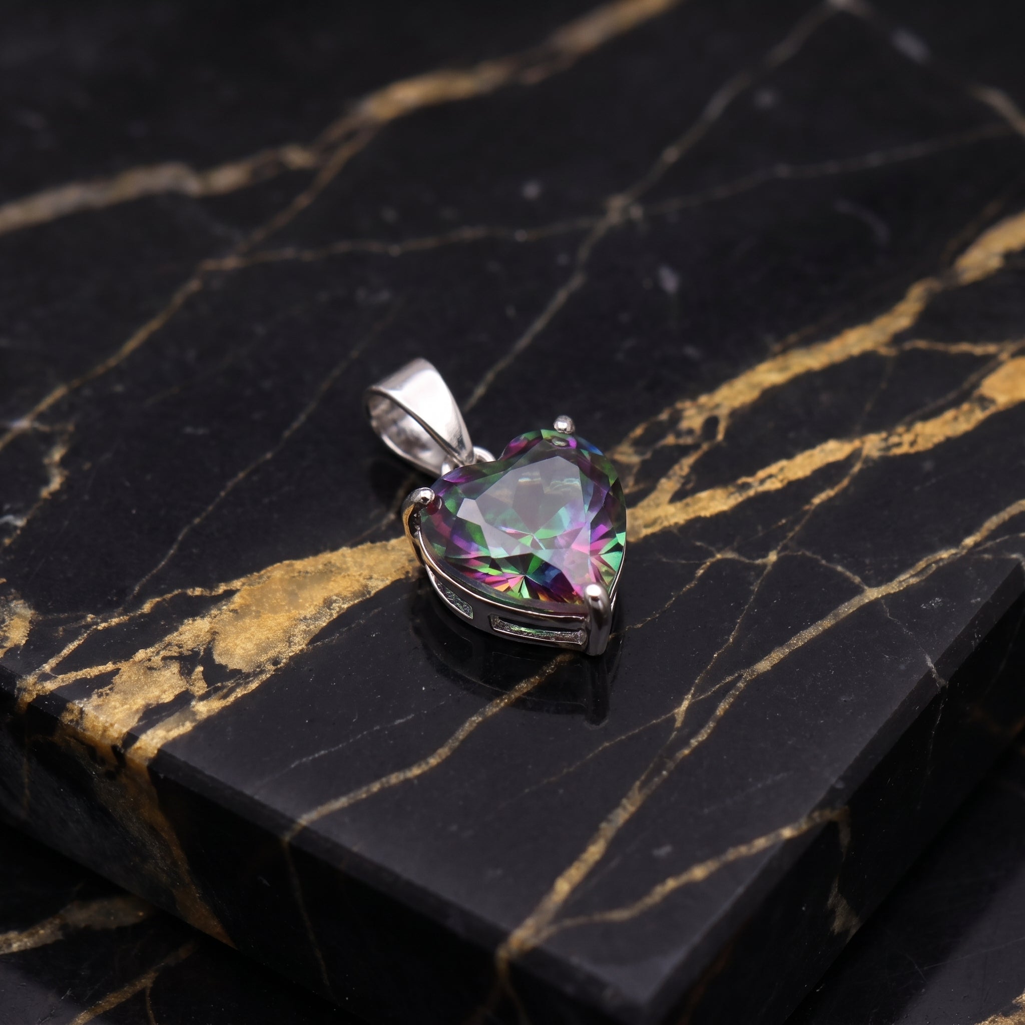 A heart-shaped iridescent gemstone pendant in a silver setting, displayed on a polished black and gold marble slab.