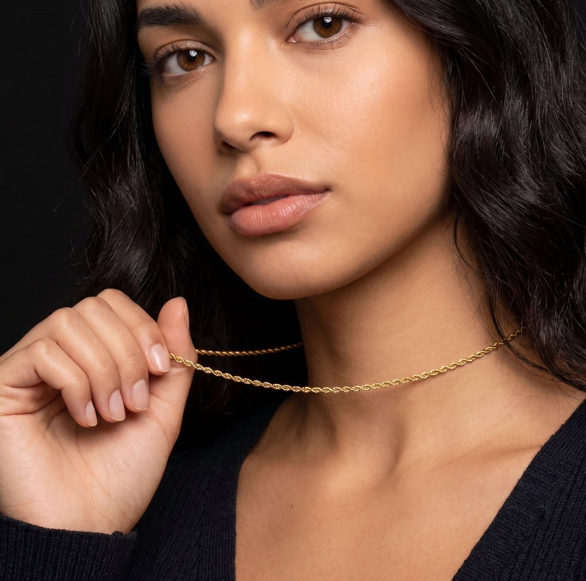 A close-up of a woman wearing a delicate gold chain necklace, holding one end of the chain with her hand against a dark background.