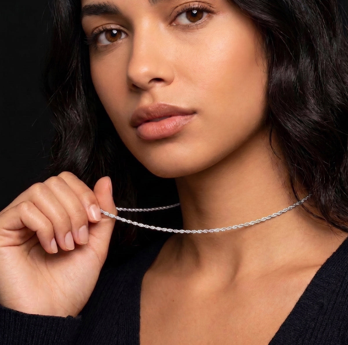A close-up of a woman wearing a delicate silver chain necklace, holding one end of the chain with her hand against a solid black background.