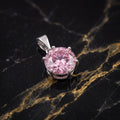 A round-cut pink gemstone pendant in a silver-toned prong setting, displayed on a black marble surface with gold veining.