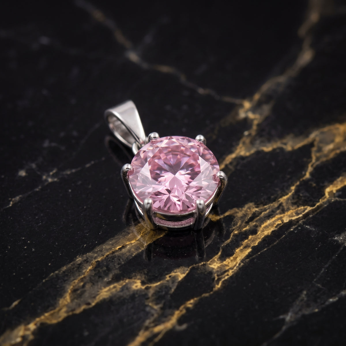 A round-cut pink gemstone pendant in a silver-toned prong setting, displayed on a black marble surface with gold veining.